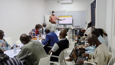 Um grupo de homens sentados em uma sala de conferências no Mali, ouvindo um homem fazer uma apresentação com uma televisão grande