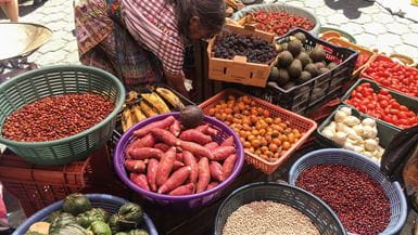 Bacias de plástico e caixotes em um mercado de rua na Guatemala, contendo diferentes tipos de feijões, grãos, frutas e legumes coloridos