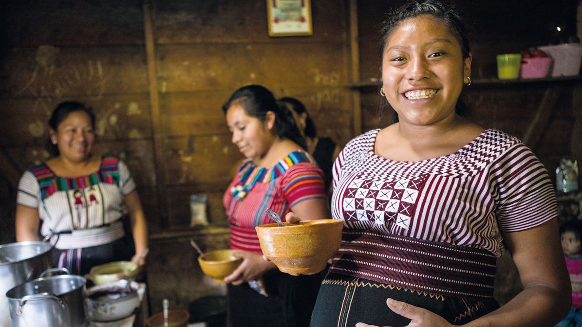 Tres mujeres guatemaltecas sonrientes, una de ellas en estado avanzado de embarazo, sostienen tazones de comida en una cocina con paredes de madera.