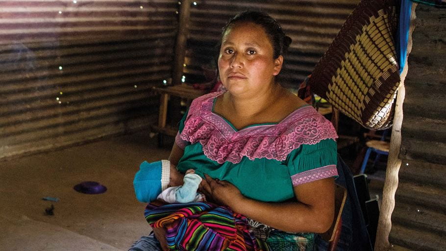 Una madre guatemalteca amamanta a su bebé en una habitación con paredes de láminas de zinc.