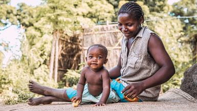 Uma mãe do Malawi sentada em uma esteira sorrindo para a filha que olha para a câmera