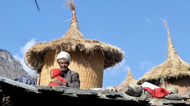 Un homme est assis avec un petit enfant sur un toit devant un grand grenier à céréales en bambou avec un toit en paille pointu 