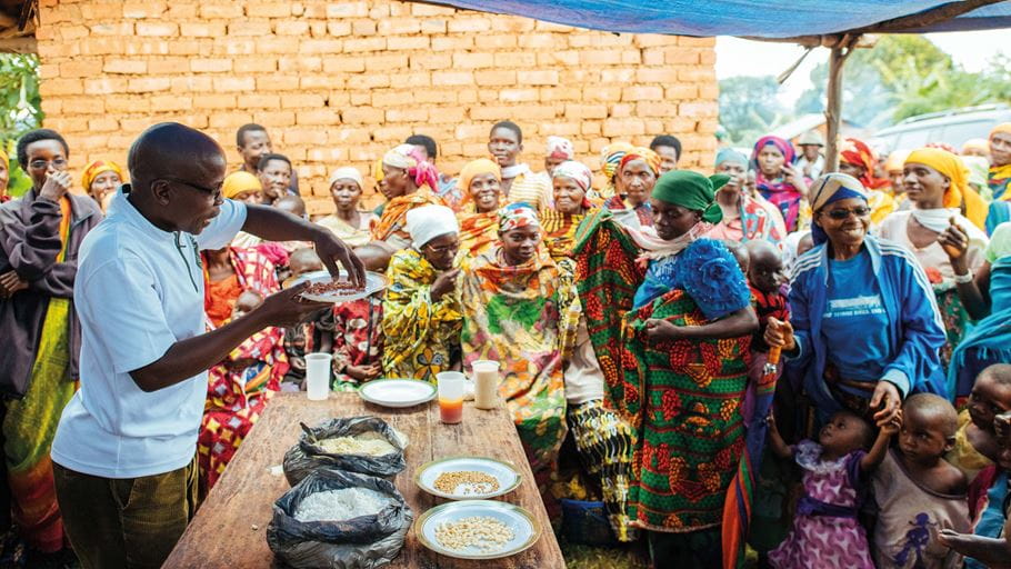 Un grupo de mujeres, niños y niñas observa a un hombre de Burundi que sostiene un plato de granos sobre unas bolsas y platos que contienen diferentes tipos de harina, frijoles y granos colocados sobre una mesa de madera.