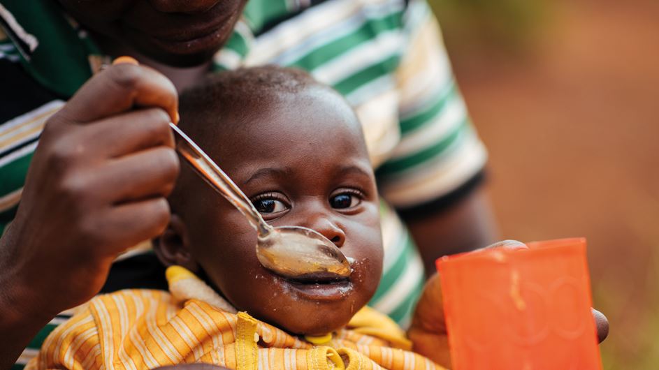 Uma criança no Burundi comendo mingau com uma colher prateada segurada por um adulto