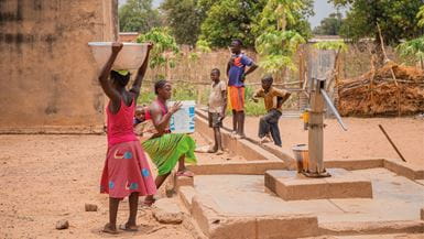A group of young people gather around a community water pump and a girl wearing a pink skirt and shirt carries a large bowl on her head