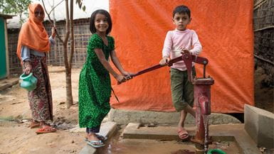 Two children use a red metal water pump in their community in Bangladesh, standing next to a woman carrying a green jug.