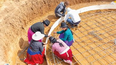 A group of men and women work at the bottom of a sandy pit, preparing the ground for a water tank
