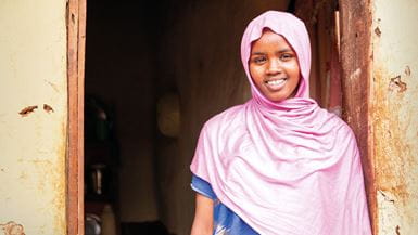 Uma jovem estudante usando um véu rosa cobrindo a cabeça, sorrindo sob o umbral de uma porta na Etiópia