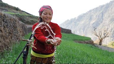 A Nepali woman stands on a green rice paddy with hills in the background and washes her hands under a water pipe.