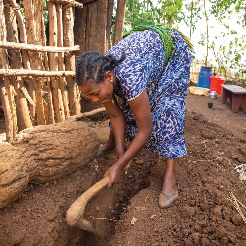 A woman, wearing a blue patterned dress, digs a trench in the soil using a wooden tool for a water pipe in rural Ethiopia.