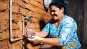 A smiling Brazilian woman collects water from a running tap fixed to a red brick wall.