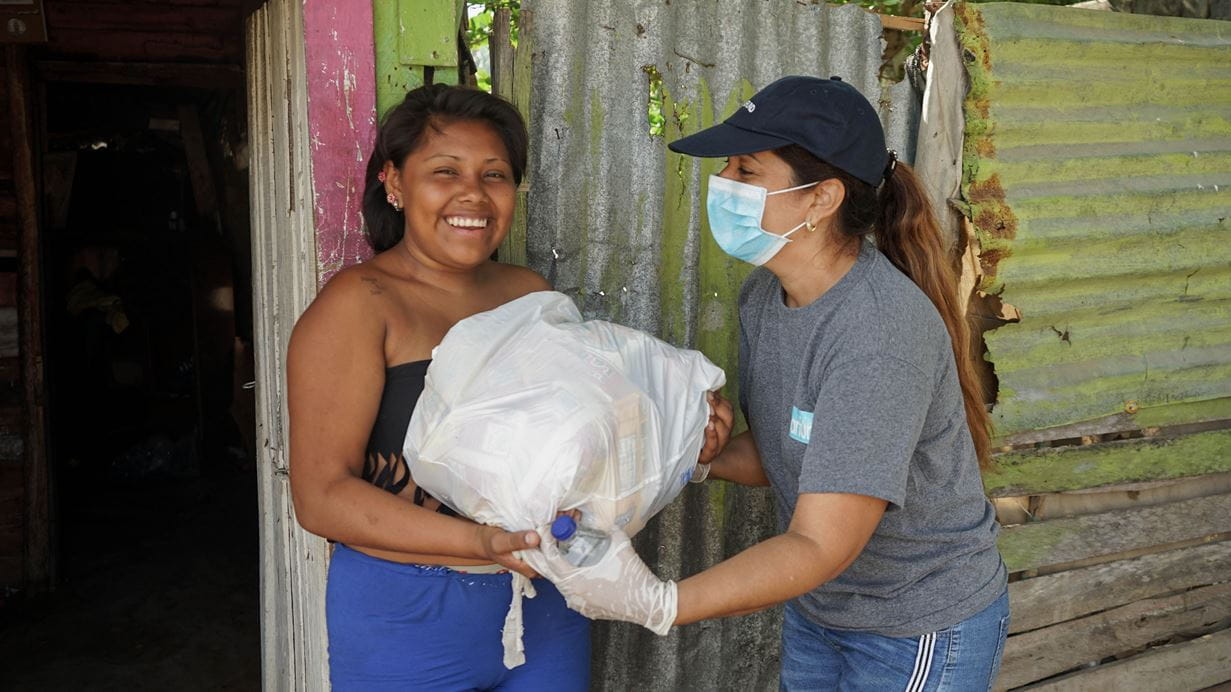 A lady with a face-mask giving a food parcel to a smiling woman in Columbia