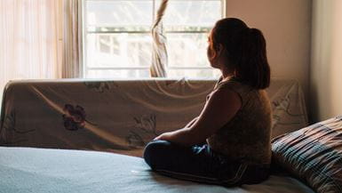 A young girl sitting on a bed silhouetted against a window