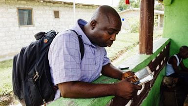 A Liberian man wearing a backpack leaning on a counter and checking his phone