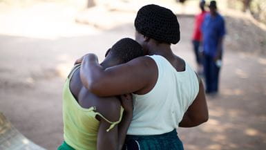 A motherly Malawian woman stands ini a rural village with her arm around the shoulders of a younger woman, both with their backs to the camera.