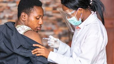 An African man receiving a vaccination on his shoulder from a nurse wearing protective equipment.