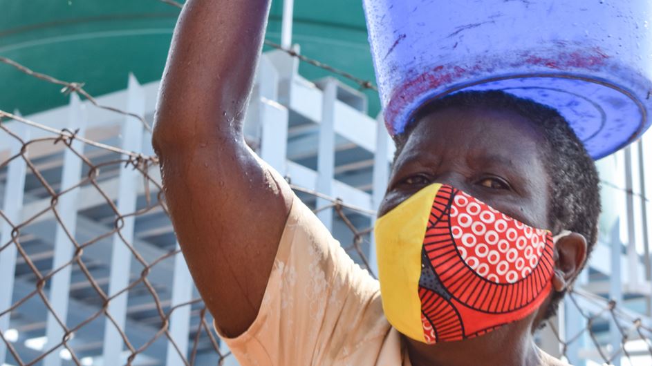 A Zimbabwean woman wearing a facemask holding a bucket of water on her head.