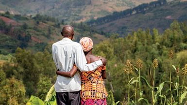 A husband and wife in Rwanda stand with their arms around each other and look out over a view of trees and hills