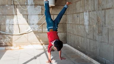 A young boy dressed in blue jeans and a red t-shirt does a handstand next to a house in Lebanon