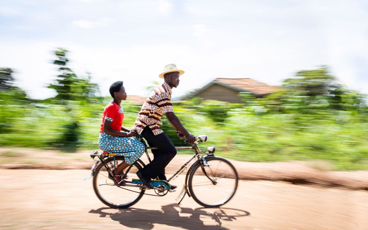 Un homme rwandais à vélo, coiffé d’un chapeau et vêtu d’une chemisette bariolée, transporte sur le porte-bagage arrière une femme rwandaise vêtue d’habits colorés.