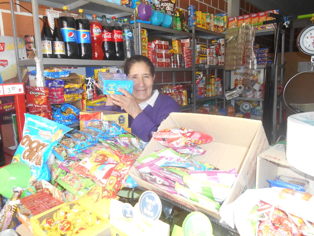 Uma mulher boliviana com um suéter roxo sorrindo para a câmera de dentro de uma mercearia bem abastecida.