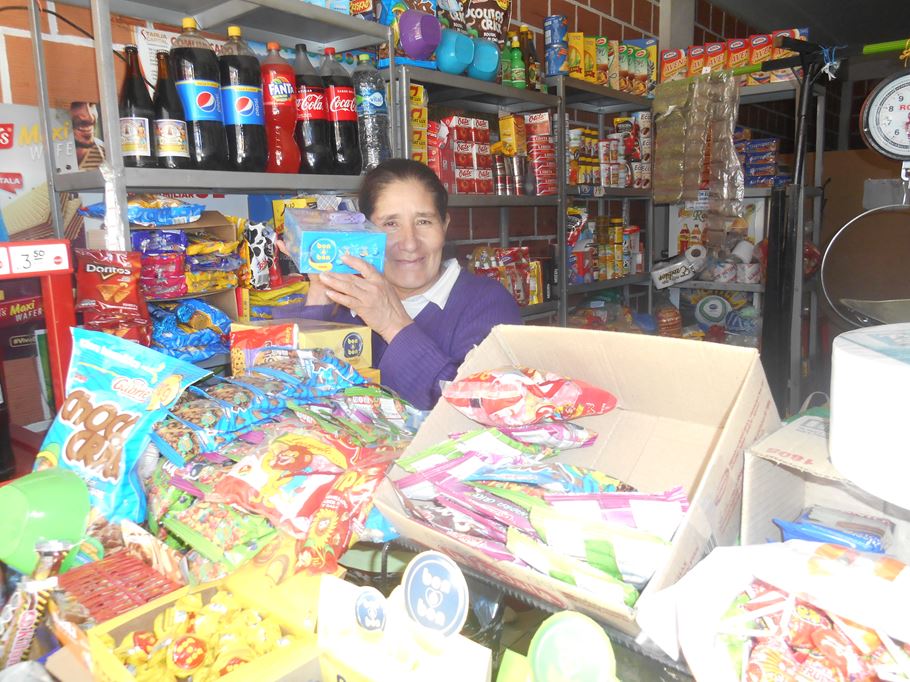 Uma mulher boliviana com um suéter roxo sorrindo para a câmera de dentro de uma mercearia bem abastecida.