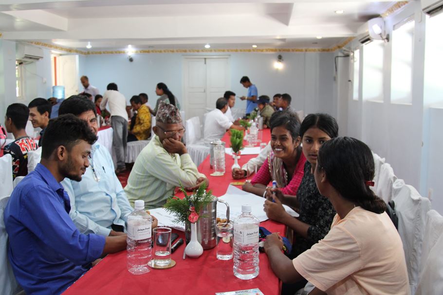 Um grande grupo de homens e mulheres nepaleses sentados ao redor de mesas dentro de um prédio, conversando e sorrindo.