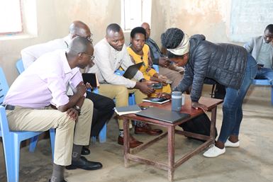 Au Rwanda, un groupe d’hommes et de femmes, assis dans une pièce, regardent un téléphone portable posé sur une table basse qu’une femme penchée en face d’eux est en train d’utiliser.