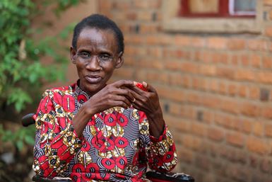 Une femme rwandaise vêtue d’un haut coloré est assise en extérieur dans un fauteuil roulant, devant le mur en briques d’un bâtiment.