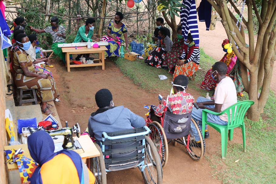 Um grupo de homens e mulheres de Ruanda sentado do lado de fora em um círculo, alguns em cadeiras de rodas, e uma pessoa com uma máquina de costura em uma mesa à sua frente.