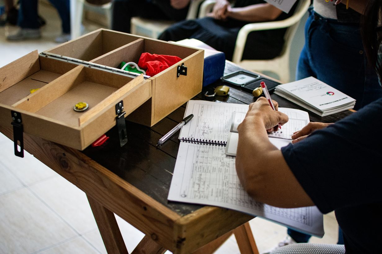 A Venezuelan woman sits at a table and writes in a record book with a wooden cash box in the background.