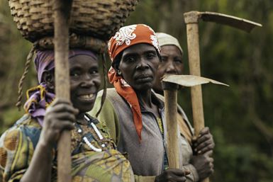 Trois agricultrices rwandaises, l’une avec un panier posé sur sa tête, se tiennent côte à côte avec des binettes à la main, le visage tourné vers l’objectif.
