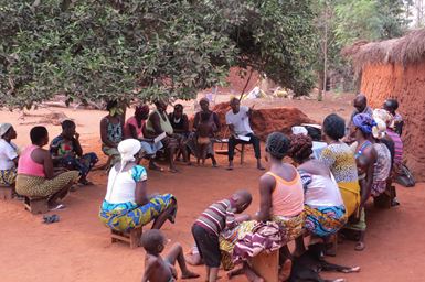 Un grand groupe de femmes, d’hommes et d’enfants togolais sont assis sur des bancs en bois sous un arbre, à côté de maisons en terre rouge et aux toits de chaume.
