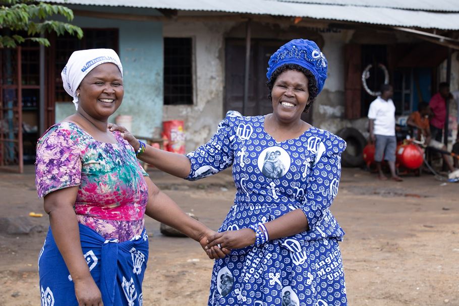 Deux femmes tanzaniennes, vêtues essentiellement de tenues et de coiffes bleues et blanches, se saluent chaleureusement dans la rue.