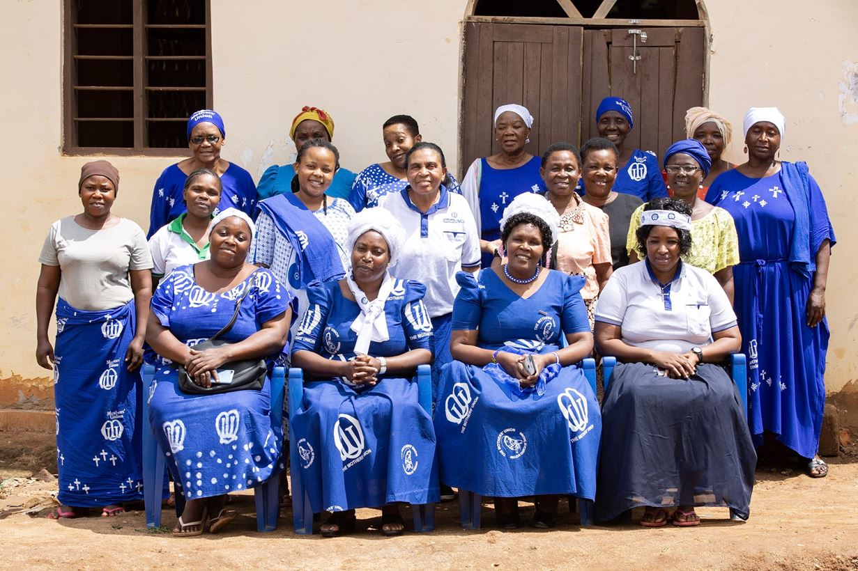 Un groupe de femmes tanzaniennes, vêtues de tenues bleues et blanches, posent face à l’objectif devant un bâtiment aux murs couleur crème orné d’une porte et d’une fenêtre en bois.
