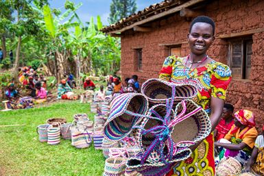 Une femme burundaise souriante, vêtue d’une robe bariolée, tient des paniers artisanaux, avec d’autres artisans en arrière-plan.