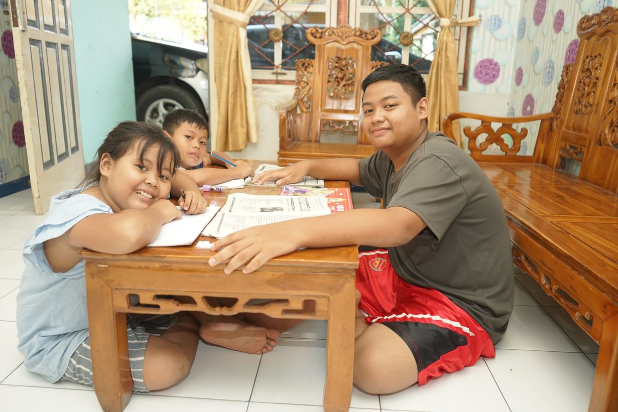 Two school children in an Indonesian home sit on the floor and lean on a table, writing on their school books, and a young man sits with them with a workbook in front of him.