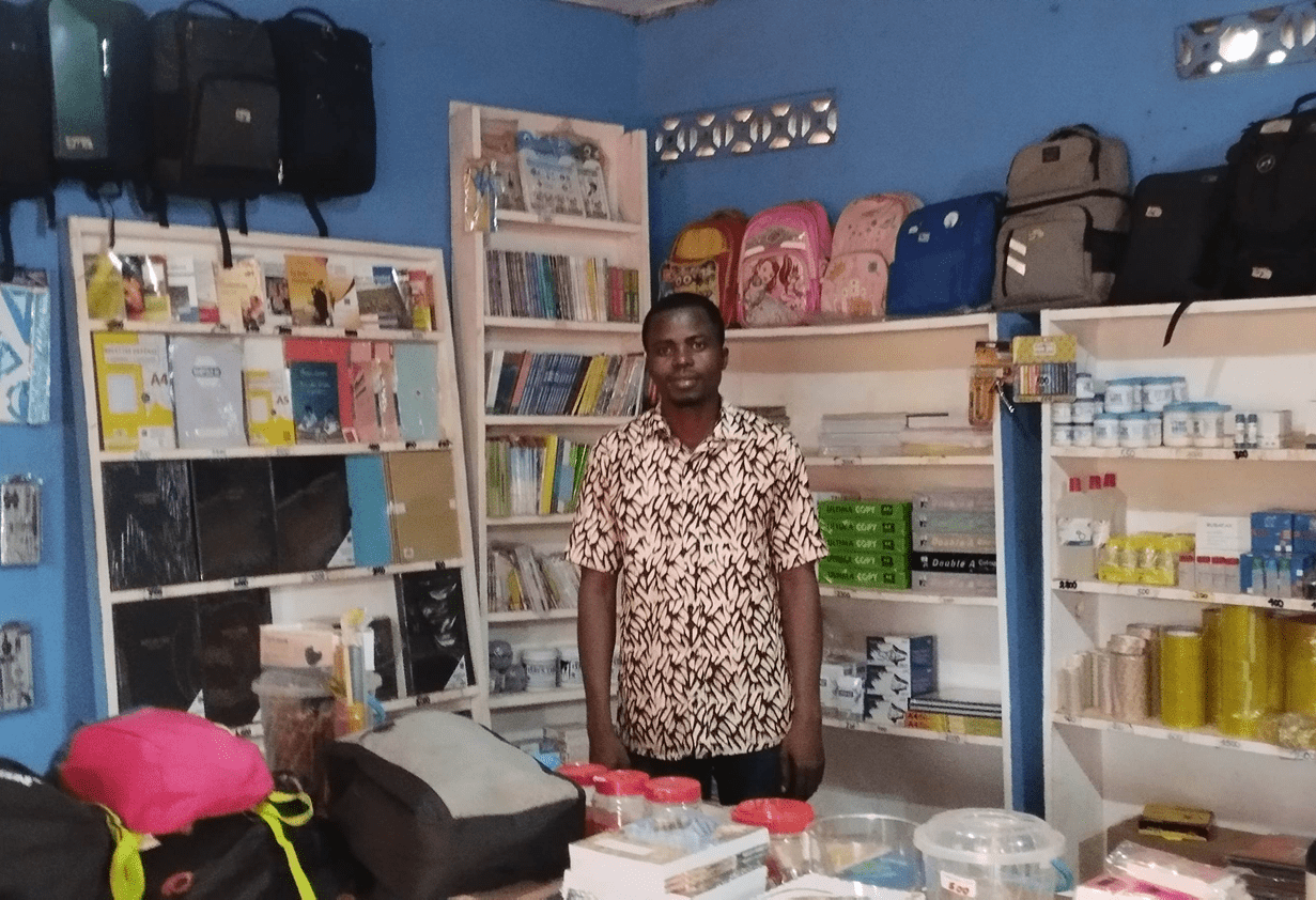 A Togolese man wearing a pink and black patterned shirt stands in his well-stocked school and office supply shop.