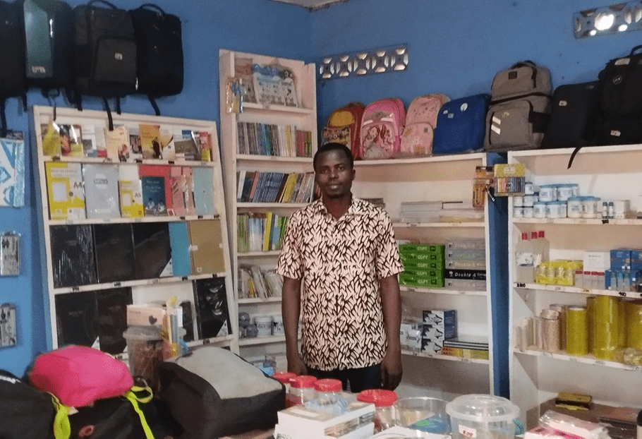 A Togolese man wearing a pink and black patterned shirt stands in his well-stocked school and office supply shop.