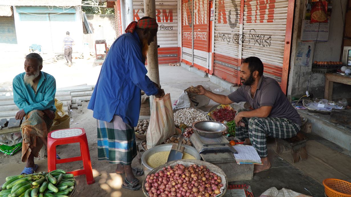 A man sits outside his shop in Bangladesh, surrounded by bowls of grains and vegetables, and hands an item of produce to a male customer.