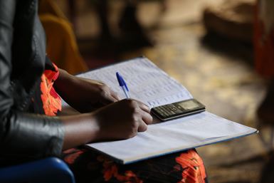 A lady neatly writes the details of financial transactions in a notebook, and uses her small mobile phone as a calculator.