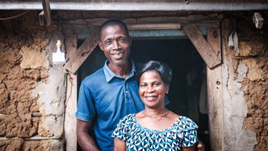 A husband and wife in Côte d’Ivoire stand in the doorway of their home and smile at the camera.