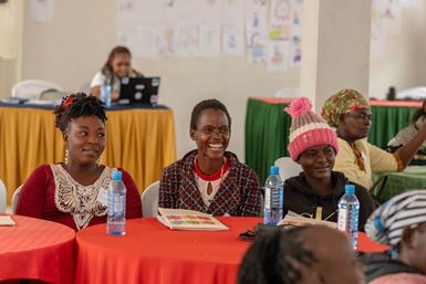 Three smiling Kenyan young people sit at a table in a room with several other people.