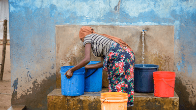 Woman fills up large, blue and red plastic buckets with water at a communal water source