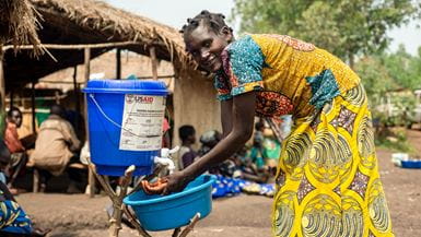 Woman from Uganda, wearing yellow patterned dress, washes hands at plastic blue water bucket and smiles at the camera.