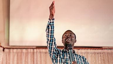 A pastor wearing a blue and white checkered, collared shirt raises his right hand in the air and talks enthusiastically into a microphone.