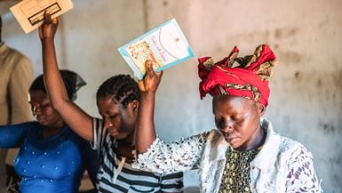 Tres mujeres con tocados coloridos sostienen folletos en el aire con las manos derechas y cierran los ojos durante un servicio en Malawi.