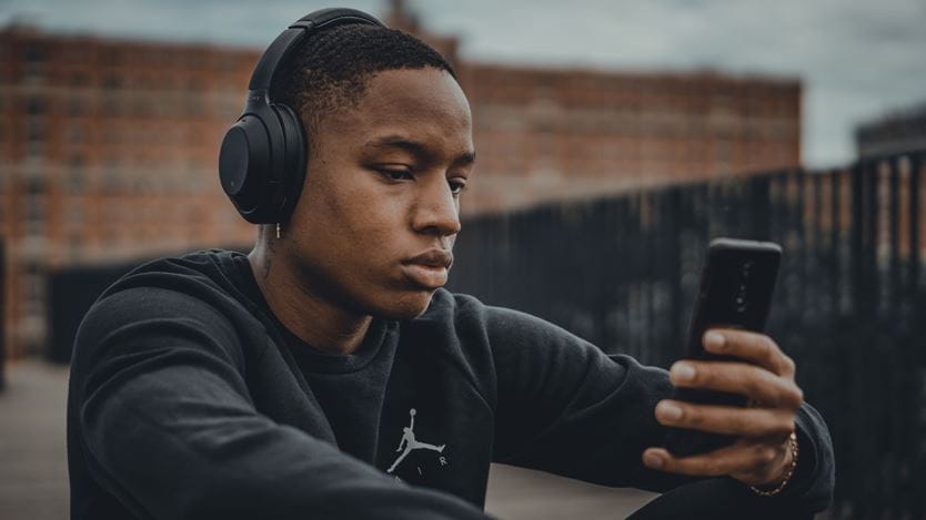 Man wearing black jumper and back heaphones holds phone in hands