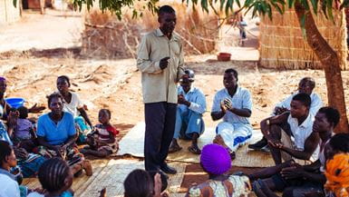 Un chef communautaire au Malawi se tient au milieu d’un groupe assis en cercle pour discuter de techniques de travail en commun.