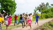 A group of children are running and playing with paper airplanes on a dirt path outdoors.